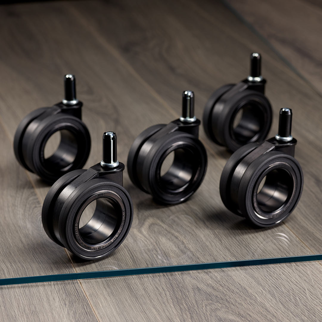 Five black hubless caster wheels for an office chair, arranged on a glass chair mat placed on a wooden floor. Each wheel has a metallic stem and a rubber tire.