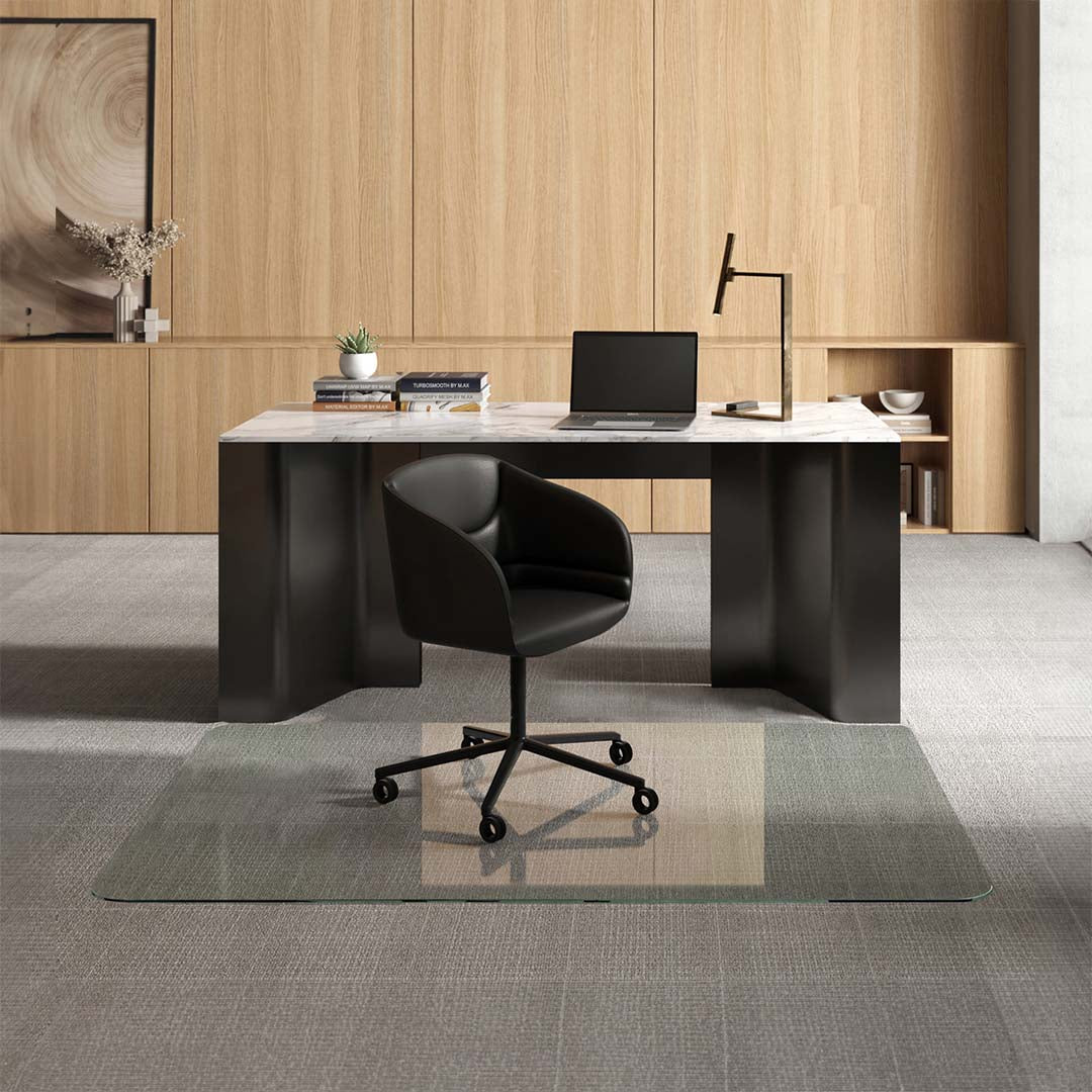 A modern office setup with a sleek black desk featuring a marble top, on which there is a laptop, several books, a small potted plant, and a desk lamp. In front of the desk, a black swivel chair is positioned on a 48" x 72" Chiaro XL glass chair mat placed over a light gray carpet. The background consists of light wooden paneling with built-in shelving displaying decorative items and more books.