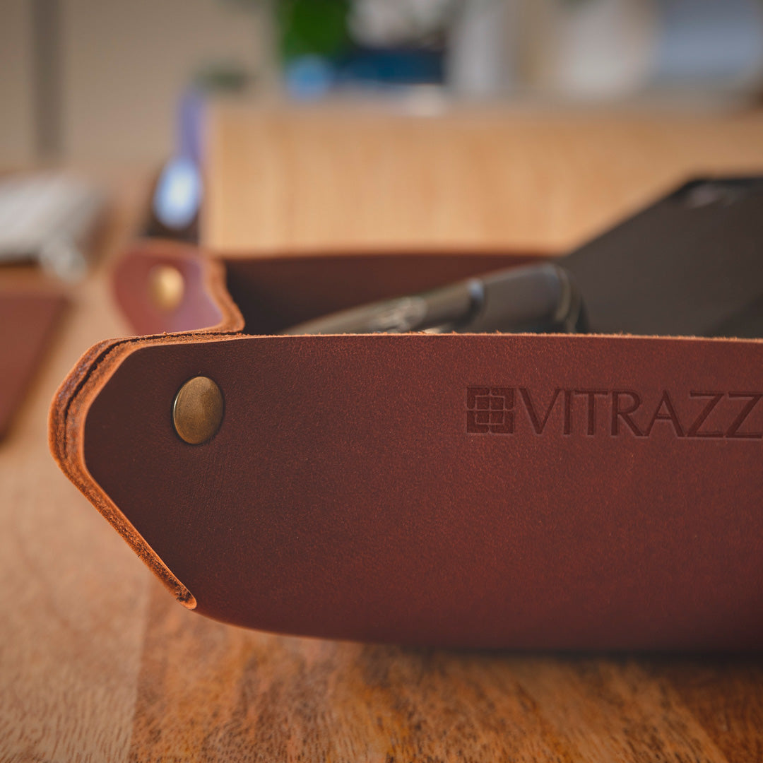 #color_mahogany A close-up image of a brown leather desk organizer with the brand name "VITRAZZA" embossed on it. The organizer is placed on a wooden desk, and inside it, there are some items, including a pen and a pair of glasses. The background is slightly blurred, showing some greenery and a light-colored wall.