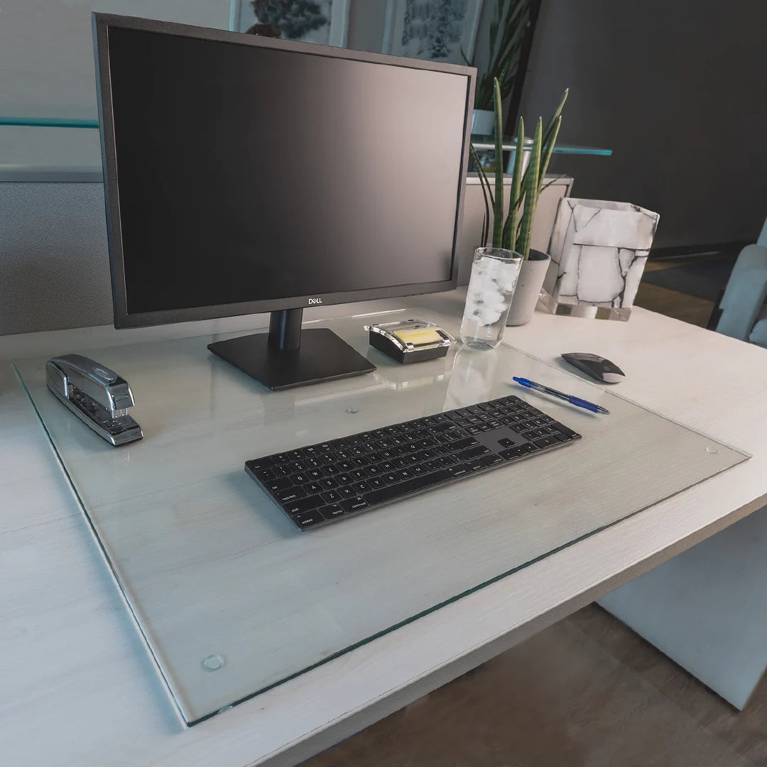 #glass clarity_chiaro (standard clear) alt="A modern office desk setup with a monitor, keyboard, mouse, stapler, glass of water with ice, pen, small plant, sticky note dispenser, and a decorative lamp on a "chiaro" glass desk blotter."