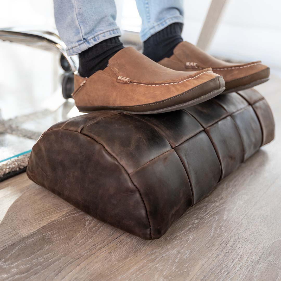 #color_espresso alt="A person wearing blue jeans, black socks, and brown leather loafers is resting their feet on a dark brown, quilted leather foot rest placed on a light wooden floor. The foot rest has a stitched pattern, giving it a cushioned appearance."