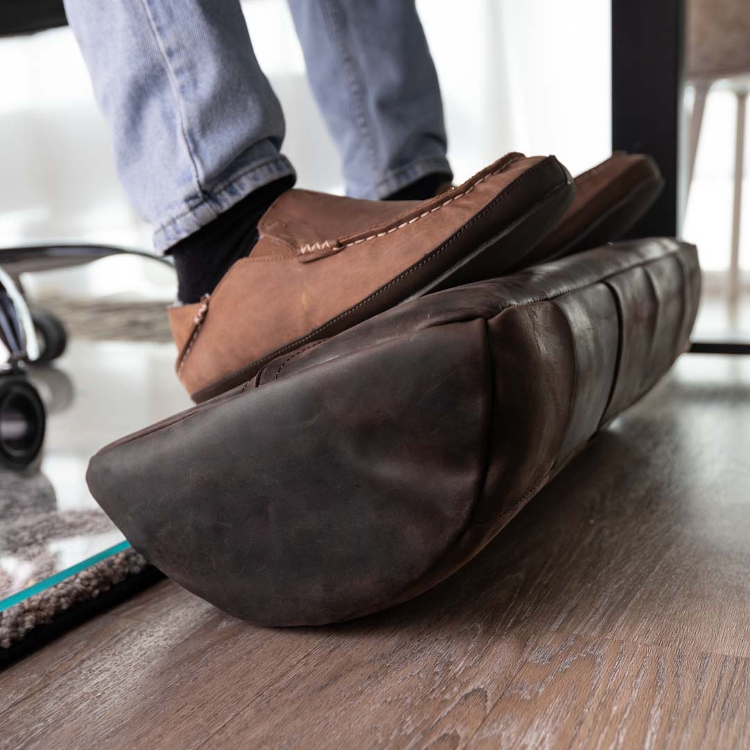 #color_espresso alt="A person wearing blue jeans and brown leather loafers is resting their feet on a dark brown, quilted leather foot rest. The foot rest is placed on a light wooden floor, and part of a glass chair mat is visible in the background."