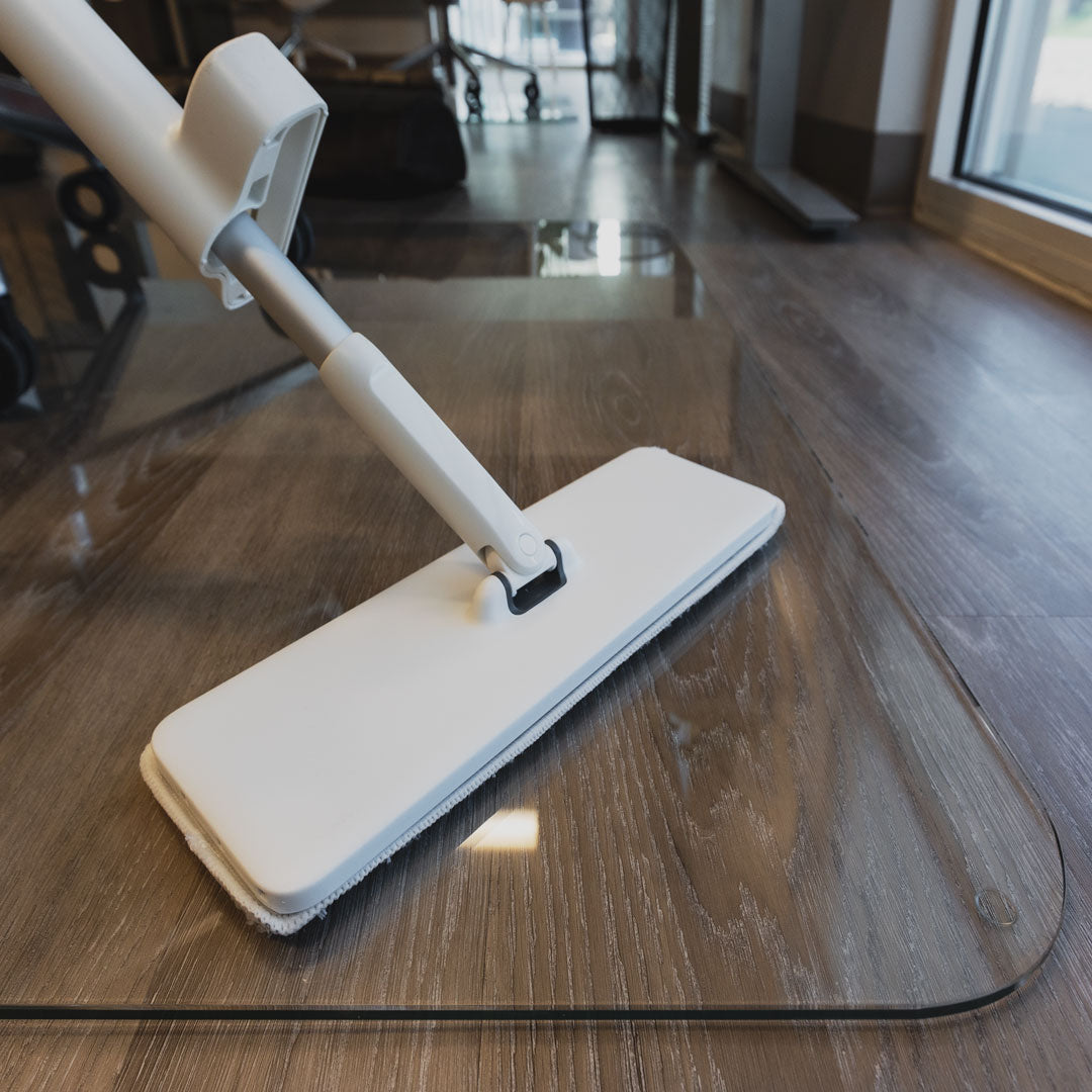 #option_mod mop alt="A white mop being used on a glass office chair mat. The mop head is flat and rectangular, with a white cloth attached. In the background, there are blurred elements of an indoor space, including furniture and large windows."