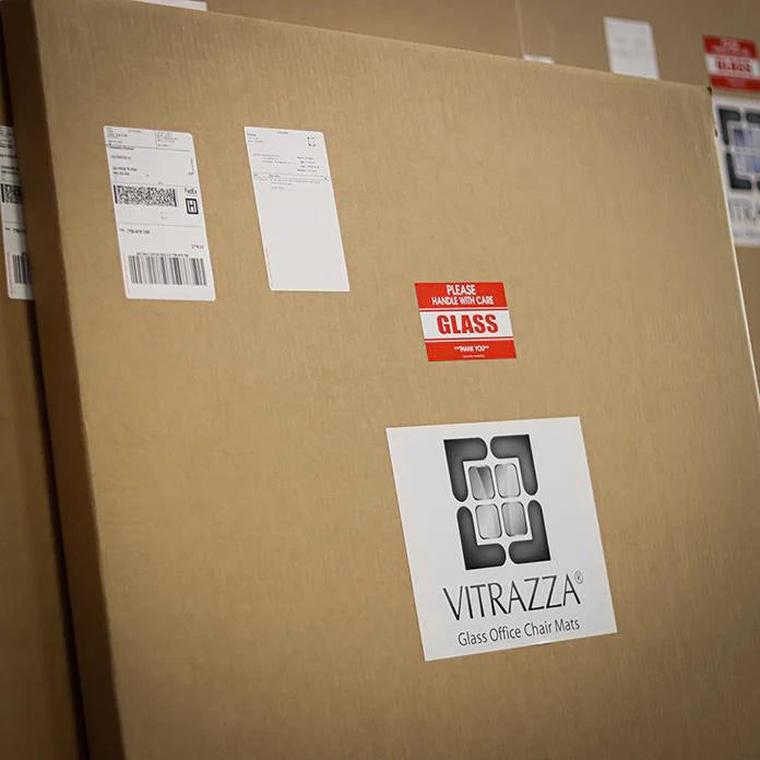 Cardboard box containing a Vitrazza glass chair mat, packaged and ready for shipment. The box is labeled with shipping details, a 'Please Handle with Care, Glass' sticker, and the Vitrazza logo.