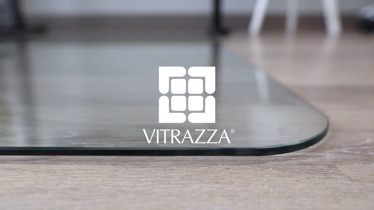 Close-up of a glass chair mat with rounded corners placed on a wooden floor. The glass image the "VITRAZZA®" logo, which consists of a stylized geometric design resembling a maze or interconnected squares, with the brand name written below it in white text. The background is slightly blurred, showing parts of furniture legs.
