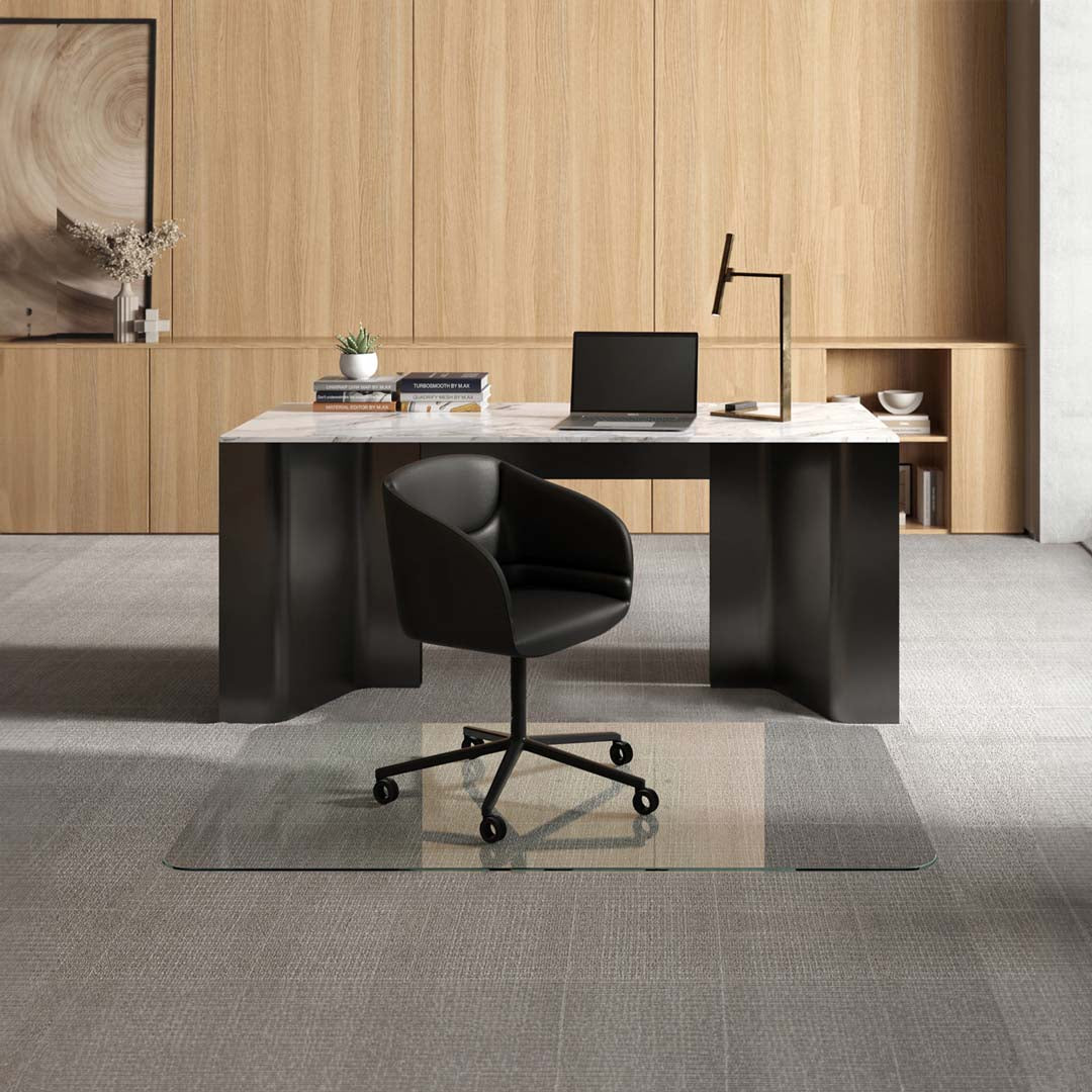 #glass clarity_luminoso (low-iron ultra clear) alt="A modern office setup with a sleek black desk featuring a marble top, on which there is a laptop, several books, a small potted plant, and a desk lamp. In front of the desk, a black swivel chair is positioned on a 42" x 64" luminoso XL glass chair mat placed over a light gray carpet. The background consists of light wooden paneling with built-in shelving displaying decorative items and more books."
