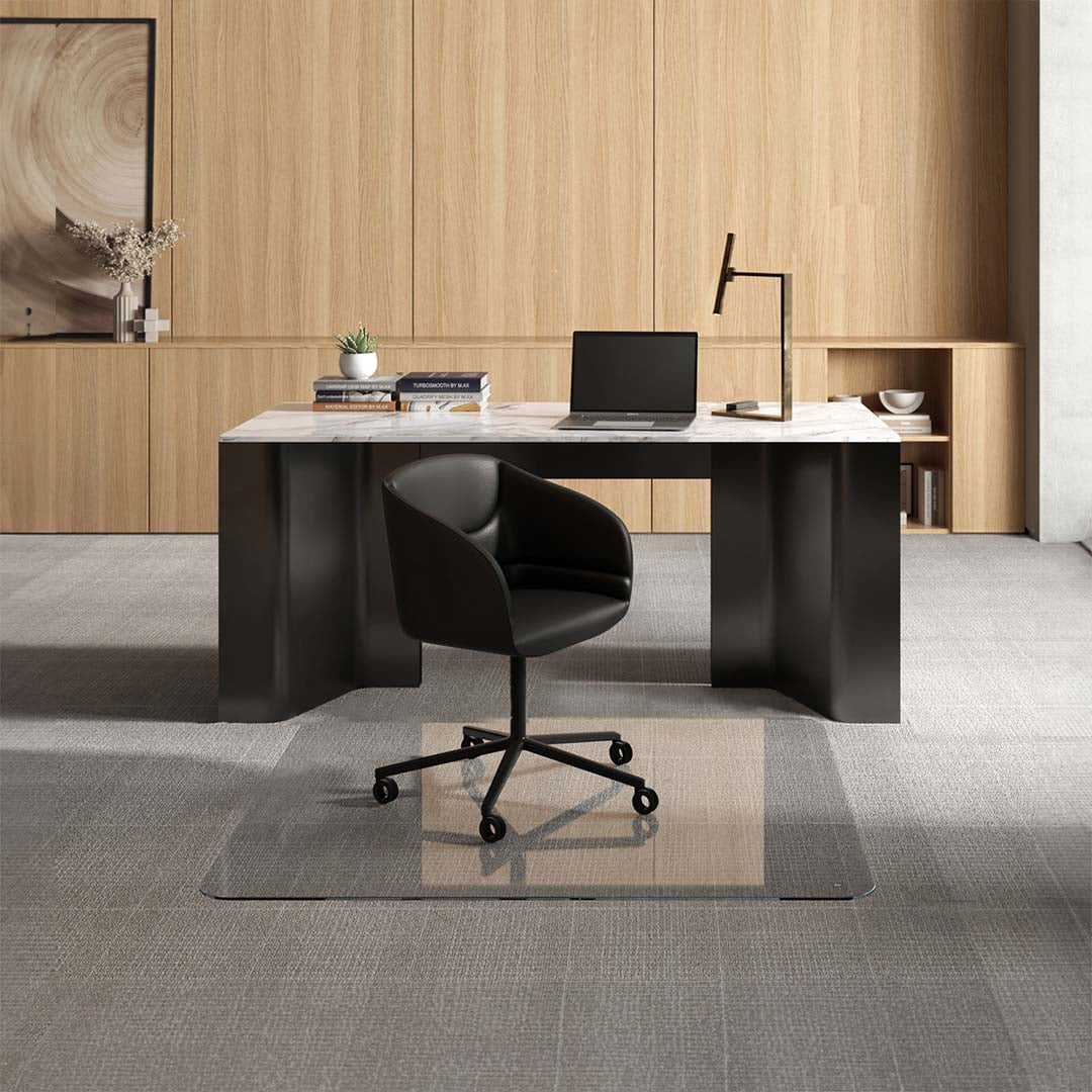 #glass clarity_luminoso (low-iron ultra clear) alt="A modern office setup with a sleek black desk featuring a marble top, on which there is a laptop, several books, a small potted plant, and a desk lamp. In front of the desk, a black swivel chair is positioned on a 54" x 54" luminoso XL glass chair mat placed over a light gray carpet. The background consists of light wooden paneling with built-in shelving displaying decorative items and more books."
