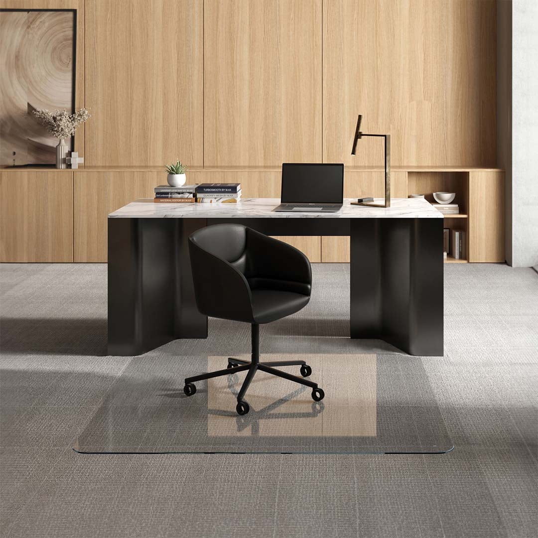 #glass clarity_luminoso (low-iron ultra clear) alt="A modern office setup with a sleek black desk featuring a marble top, on which there is a laptop, several books, a small potted plant, and a desk lamp. In front of the desk, a black swivel chair is positioned on a 60" x 60" luminoso XL glass chair mat placed over a light gray carpet. The background consists of light wooden paneling with built-in shelving displaying decorative items and more books."
