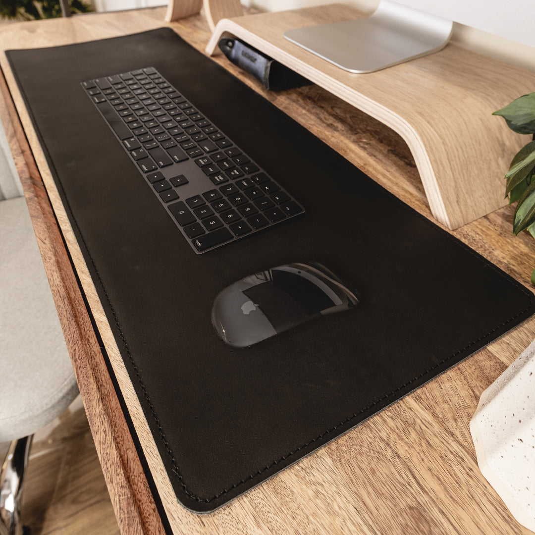 #color_charcoal A close-up view of a wooden desk setup with a large black leather and felt desk mat. On the mat, there is a black wireless keyboard and a black wireless mouse. Above the keyboard, there is a wooden monitor stand with a silver monitor placed on it. To the right, part of a green plant is visible.