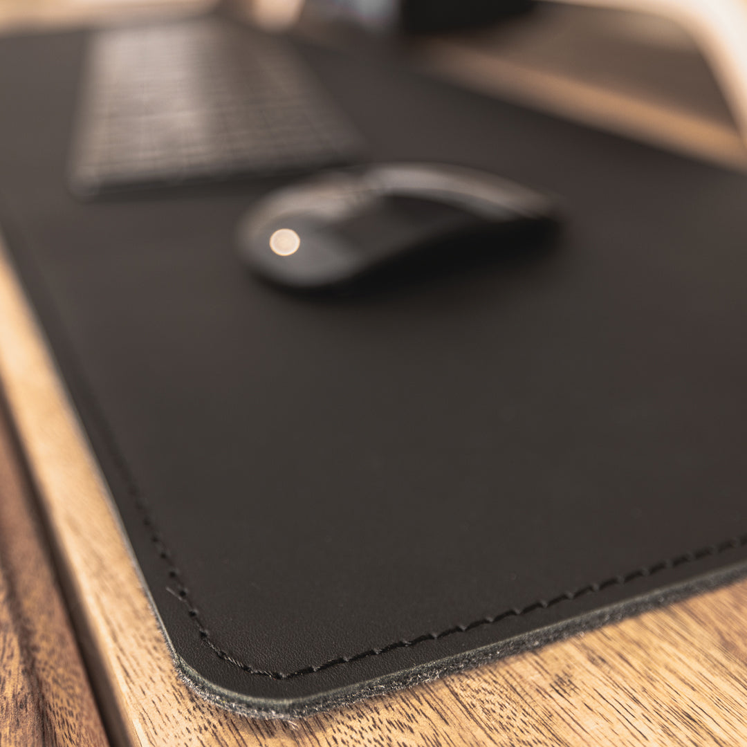 #color_charcoal A close-up image showing a portion of a wooden desk with a black leather desk mat. On the mat, there is a black wireless keyboard and a black wireless mouse. The focus is on the texture and stitching of the leather desk mat.