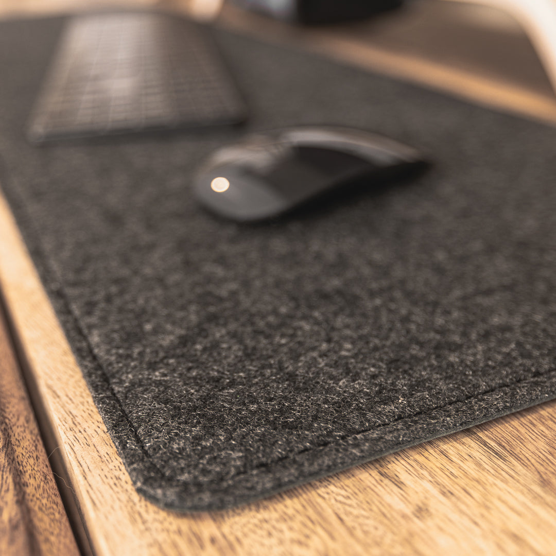 #color_charcoal A close-up image showing a portion of a wooden desk with a black felt desk mat. On the mat, there is a black wireless keyboard and a black wireless mouse. The focus is on the texture and stitching of the felt desk mat.