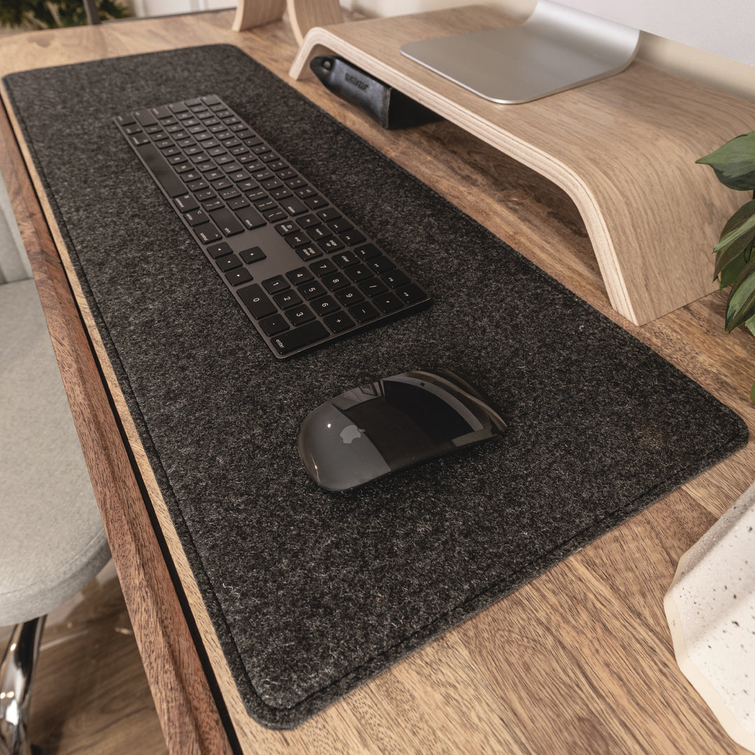 #color_charcoal A close-up view of a wooden desk setup with a large black felt desk mat. On the mat, there is a black wireless keyboard and a black wireless mouse. Above the keyboard, there is a wooden monitor stand with a silver monitor placed on it. To the right, part of a green plant is visible.