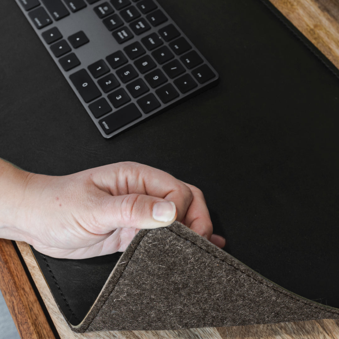 #color_charcoal A close-up of a hand lifting the corner of a black leather desk mat to reveal a felt underside. On the desk mat, there is a black wireless keyboard. The scene is set on a wooden desk.