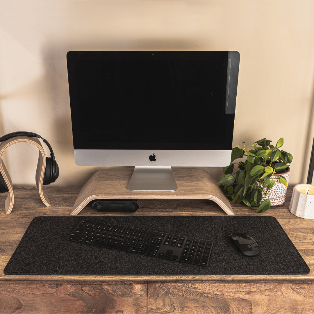#color_charcoal A minimalist office setup on a wooden desk featuring a computer monitor with a black screen, elevated on a wooden stand. In front of the monitor, there is a black felt desk mat with a black keyboard and mouse placed on it. To the left, a pair of black headphones on a wooden stand, and to the right, a potted green plant and a white candle. The background is a plain, light-colored wall.