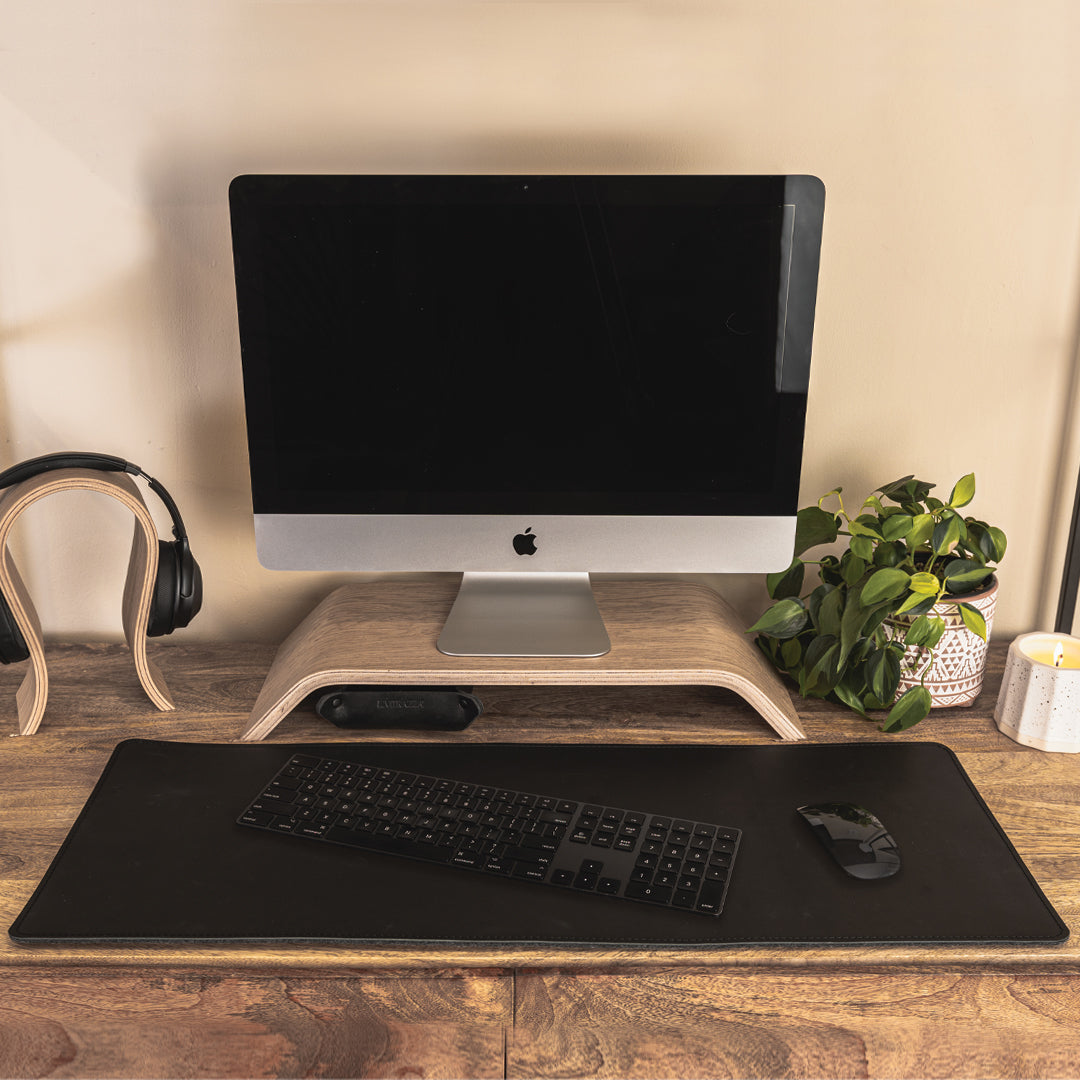 #color_charcoal A minimalist office setup on a wooden desk featuring a computer monitor with a black screen, elevated on a wooden stand. In front of the monitor, there is a black leather and felt desk mat with a black keyboard and mouse placed on it. To the left, a pair of black headphones on a wooden stand, and to the right, a potted green plant and a white candle. The background is a plain, light-colored wall.