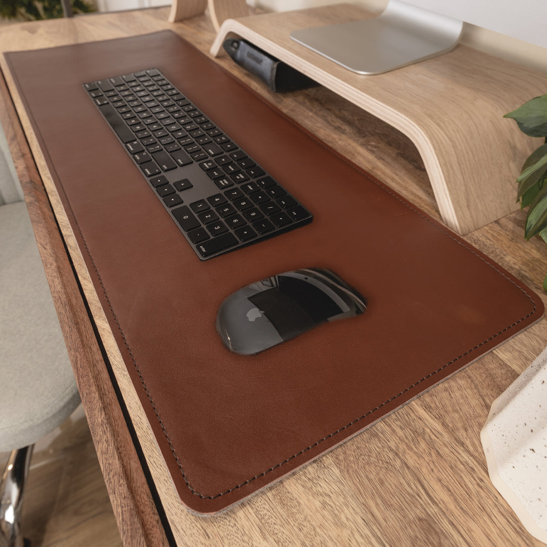 #color_mahogany A close-up view of a wooden desk setup with a large brown leather and felt desk mat. On the mat, there is a black wireless keyboard and a black wireless mouse. Above the keyboard, there is a wooden monitor stand with a silver monitor placed on it. To the right, part of a green plant is visible.