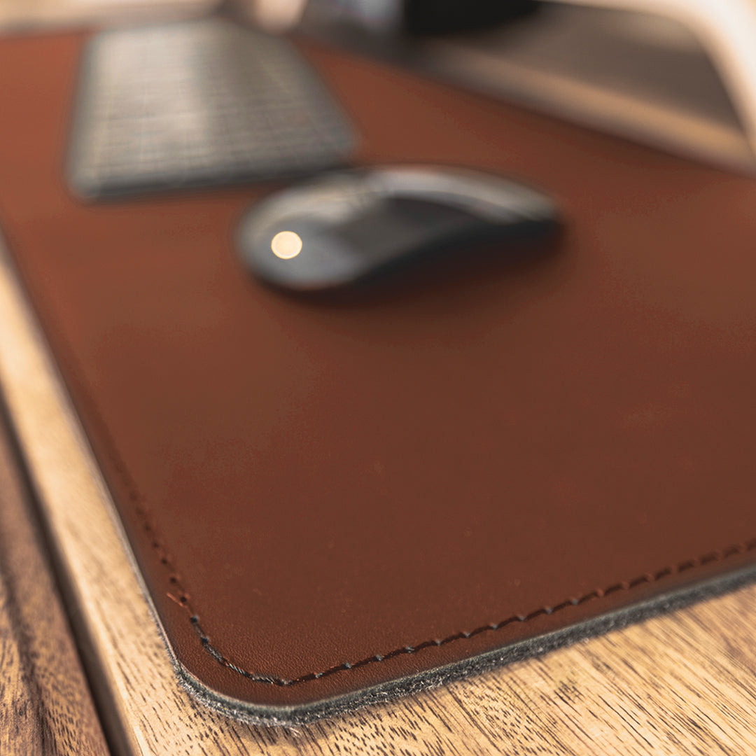 #color_mahogany A close-up image showing a portion of a wooden desk with a brown leather desk mat. On the mat, there is a black wireless keyboard and a black wireless mouse. The focus is on the texture and stitching of the leather desk mat.