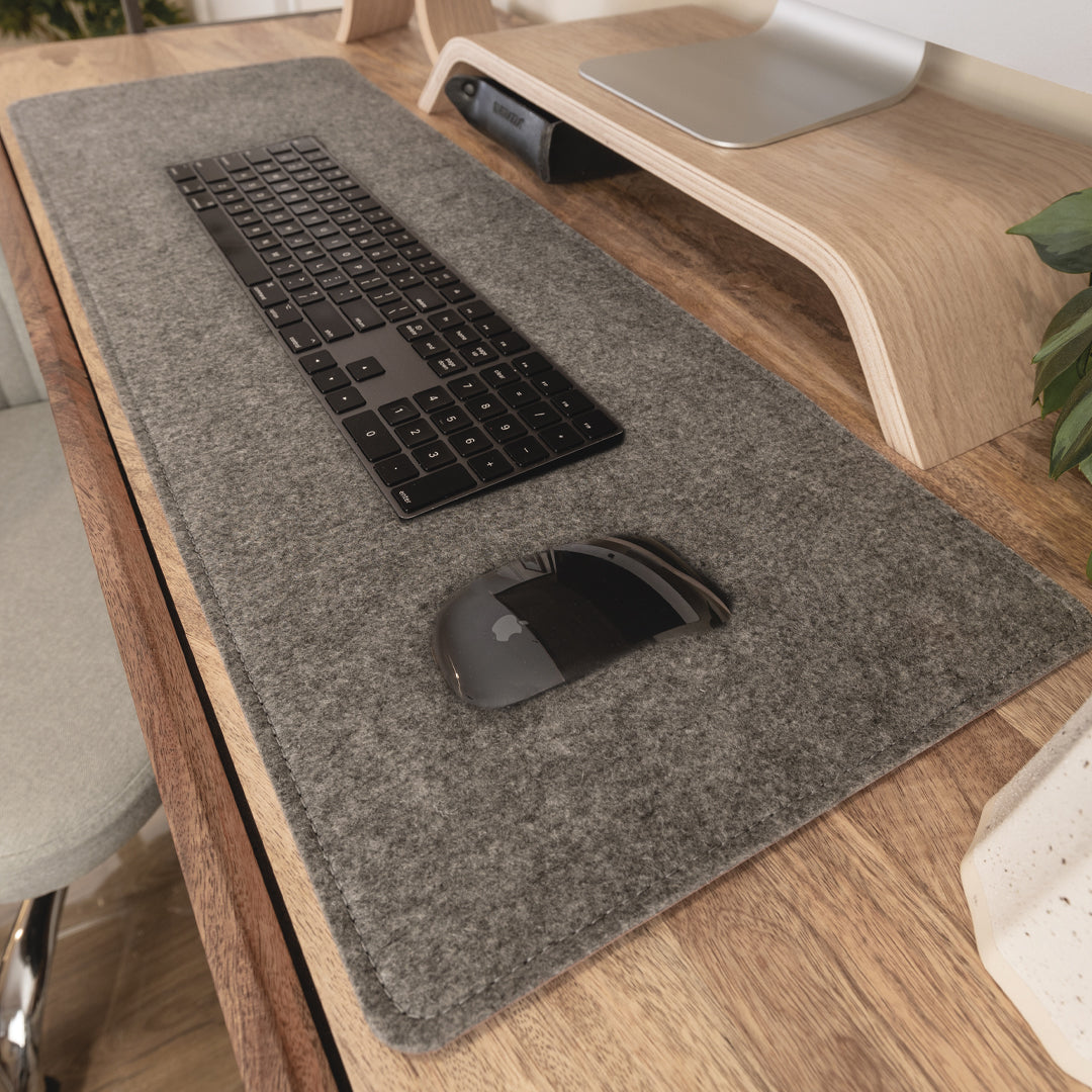 #color_mahogany A close-up view of a wooden desk setup with a large gray felt desk mat. On the mat, there is a black wireless keyboard and a black wireless mouse. Above the keyboard, there is a wooden monitor stand with a silver monitor placed on it. To the right, part of a green plant is visible.