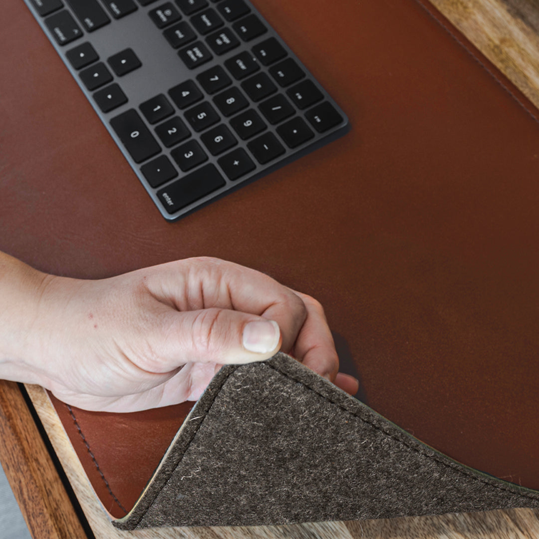 #color_mahogany A close-up of a hand lifting the corner of a brown leather desk mat to reveal a felt underside. On the desk mat, there is a black wireless keyboard. The scene is set on a wooden desk.