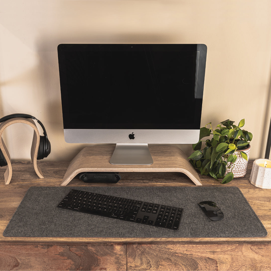 #color_mahogany A minimalist office setup on a wooden desk featuring a computer monitor with a black screen, elevated on a wooden stand. In front of the monitor, there is a grey felt desk mat with a black keyboard and mouse placed on it. To the left, a pair of black headphones on a wooden stand, and to the right, a potted green plant and a white candle. The background is a plain, light-colored wall.