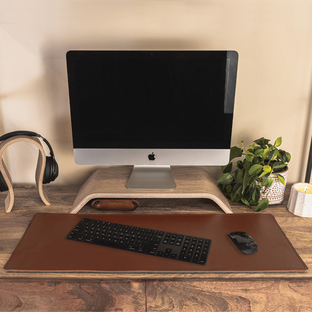 #color_mahogany A minimalist office setup on a wooden desk featuring a computer monitor with a black screen, elevated on a wooden stand. In front of the monitor, there is a brown leather and felt desk mat with a black keyboard and mouse placed on it. To the left, a pair of black headphones on a wooden stand, and to the right, a potted green plant and a white candle. The background is a plain, light-colored wall.