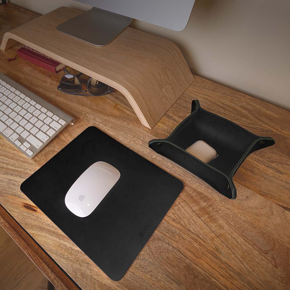 #color_charcoal alt="A minimalist wooden desk setup features a brown leather mouse pad with a white wireless mouse on it. To the left, there is a silver keyboard and a pair of sunglasses resting on a wooden monitor stand. On the right side, there is a brown leather catch-all tray holding a white ear buds case. The background includes a light-colored wall."