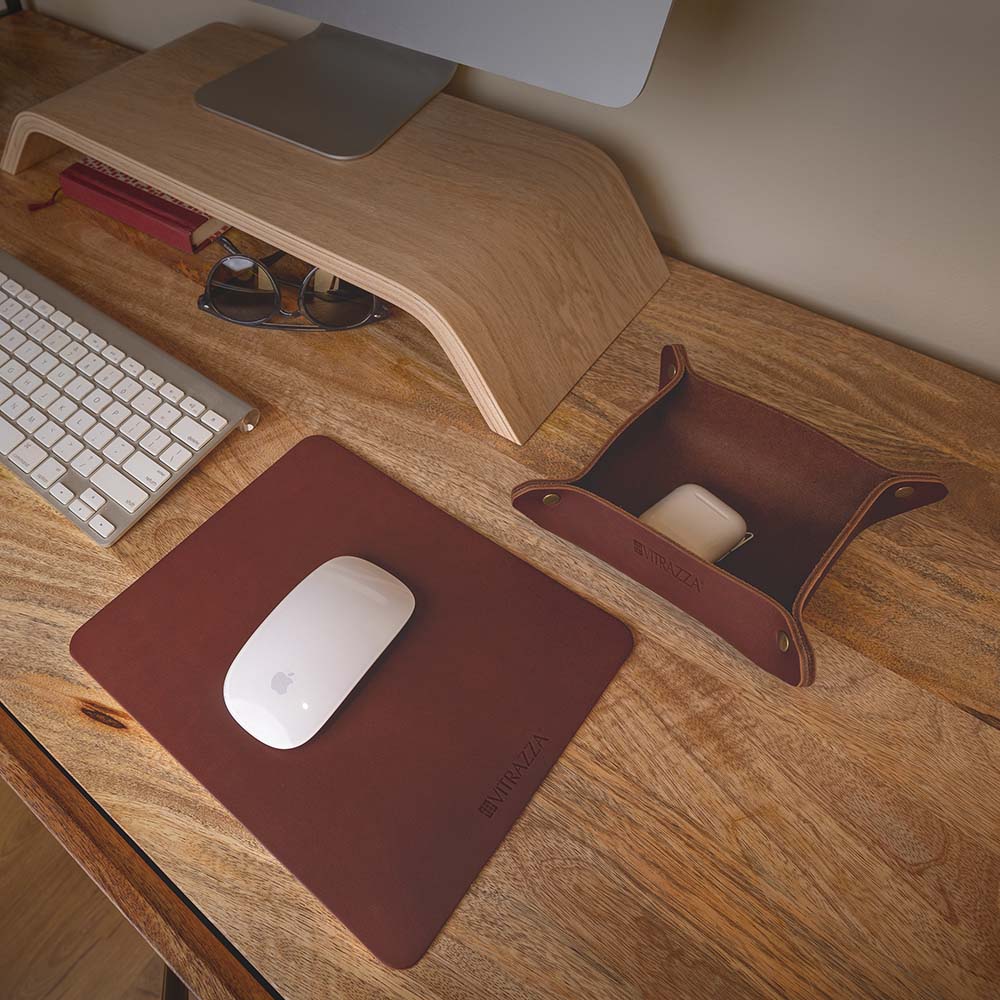 #color_mahogany alt="A minimalist wooden desk setup features a brown leather mouse pad with a white wireless mouse on it. To the left, there is a silver keyboard and a pair of sunglasses resting on a wooden monitor stand. On the right side, there is a brown leather catch-all tray holding a white ear buds case. The background includes a light-colored wall."