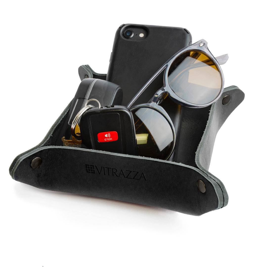 #color_charcoal alt="A black leather desk organizer with various items inside. The items include a black smartphone, a pair of sunglasses, a set of keys with a key fob that has a red button and another ear buds. The organizer is placed against a white background."
