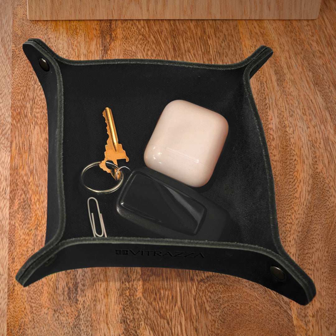 #color_charcoal alt="A black leather desk organizer is placed on a wooden desk. Inside the organizer, there are several items including a white ear buds case, a black car key fob, a set of keys with a gold key, and a silver binder clip. The background is out of focus, showing a light-colored wall and some other furniture."
