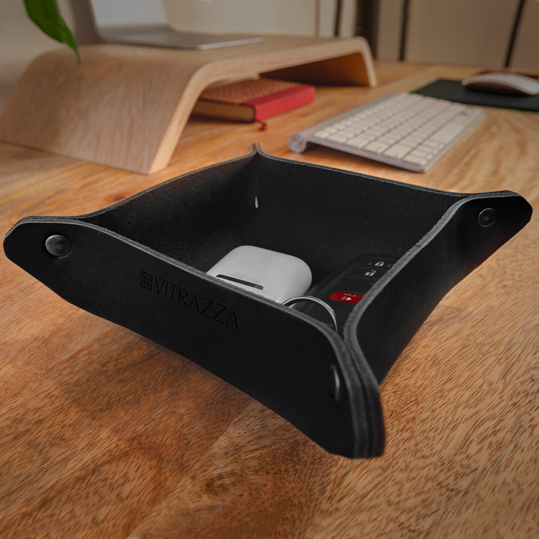 #color_charcoal alt="A black leather desk organizer is placed on a wooden desk. Inside the organizer, there are several items including a white ear buds case, a black car key fob with a red button, and a set of keys. In the background, there is a wooden monitor stand, a silver keyboard, and some books. The desk has a natural wood finish."