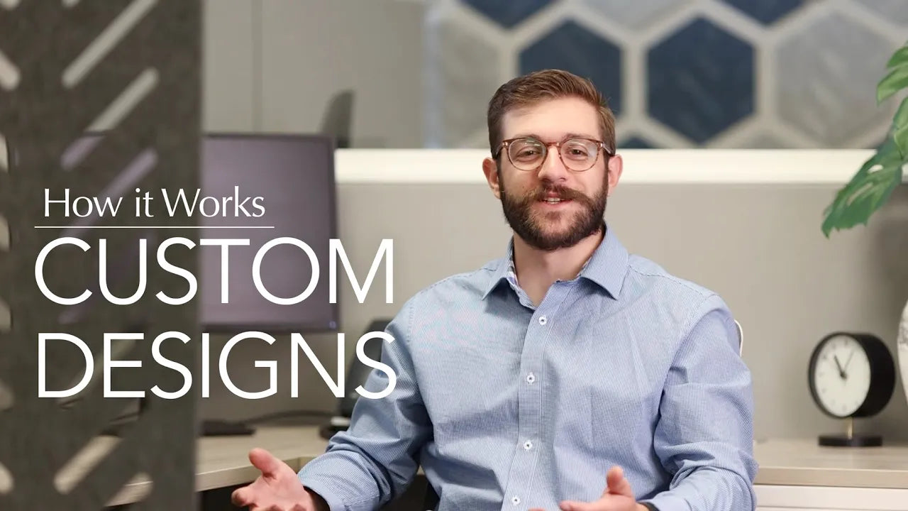 A person in a light blue button-up shirt is seated at a desk, gesturing with their hands as if explaining something. The background includes a modern office setting with a computer monitor, a plant, and a clock. The text on the image reads "How it Works CUSTOM DESIGNS".