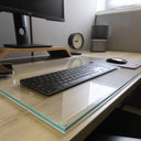 A clean and organized desk setup with a monitor on a wooden stand, a wireless keyboard on a "luminoso" glass desk blotter, a mouse on a mouse pad, a smartphone, a round analog clock, and a cylindrical speaker. The background features a light-colored wall and a window letting in natural light.#Size_18x28#Glass_Clarity_Luminoso