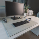 A modern office desk setup with a monitor, keyboard, mouse, stapler, glass of water with ice, pen, small plant, sticky note dispenser, and a decorative lamp on a "chiaro" glass desk blotter.#Size_24x30#Glass_Clarity_Chiaro