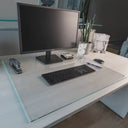 A modern office desk setup with a monitor, keyboard, mouse, stapler, glass of water with ice, pen, small plant, sticky note dispenser, and a decorative lamp on a "luminoso" glass desk blotter.#Size_24x30#Glass_Clarity_Luminoso