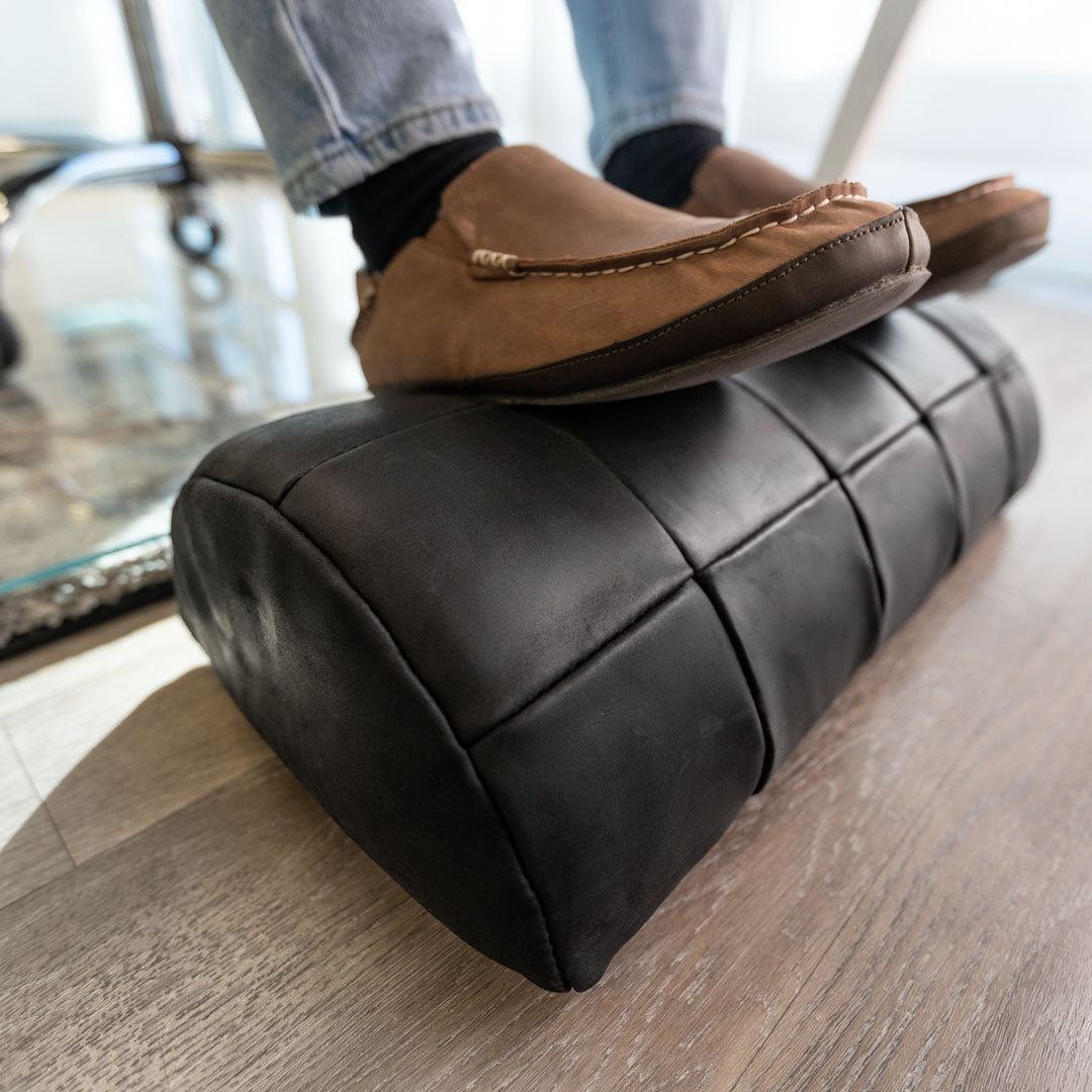 #color_charcoal alt="A person wearing blue jeans, black socks, and brown leather loafers is resting their feet on a black, quilted leather foot rest placed on a light wooden floor. The foot rest has a stitched pattern, giving it a cushioned appearance."