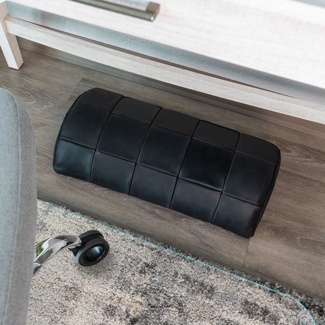 #color_charcoal alt="A black, quilted leather foot rest is placed on a light wooden floor, partially resting on a glass chair mat. In the foreground, part of an office chair wheel is visible, and there is a light gray rug with a subtle pattern underneath the chair mat. The background includes a white piece of furniture."