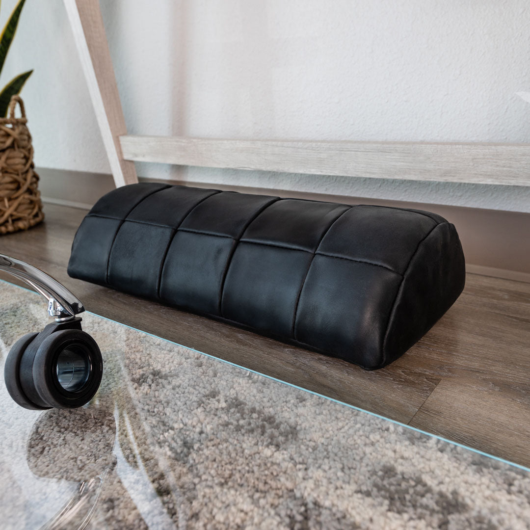 #color_charcoal alt="A black, quilted leather foot rest is placed on a light wooden floor, partially resting on a glass chair mat. In the foreground, part of an office chair wheel is visible, and there is a light gray rug with a subtle pattern underneath the chair mat. The background includes a white wall and a piece of light-colored furniture. A plant in a woven basket is also partially visible on the left side."