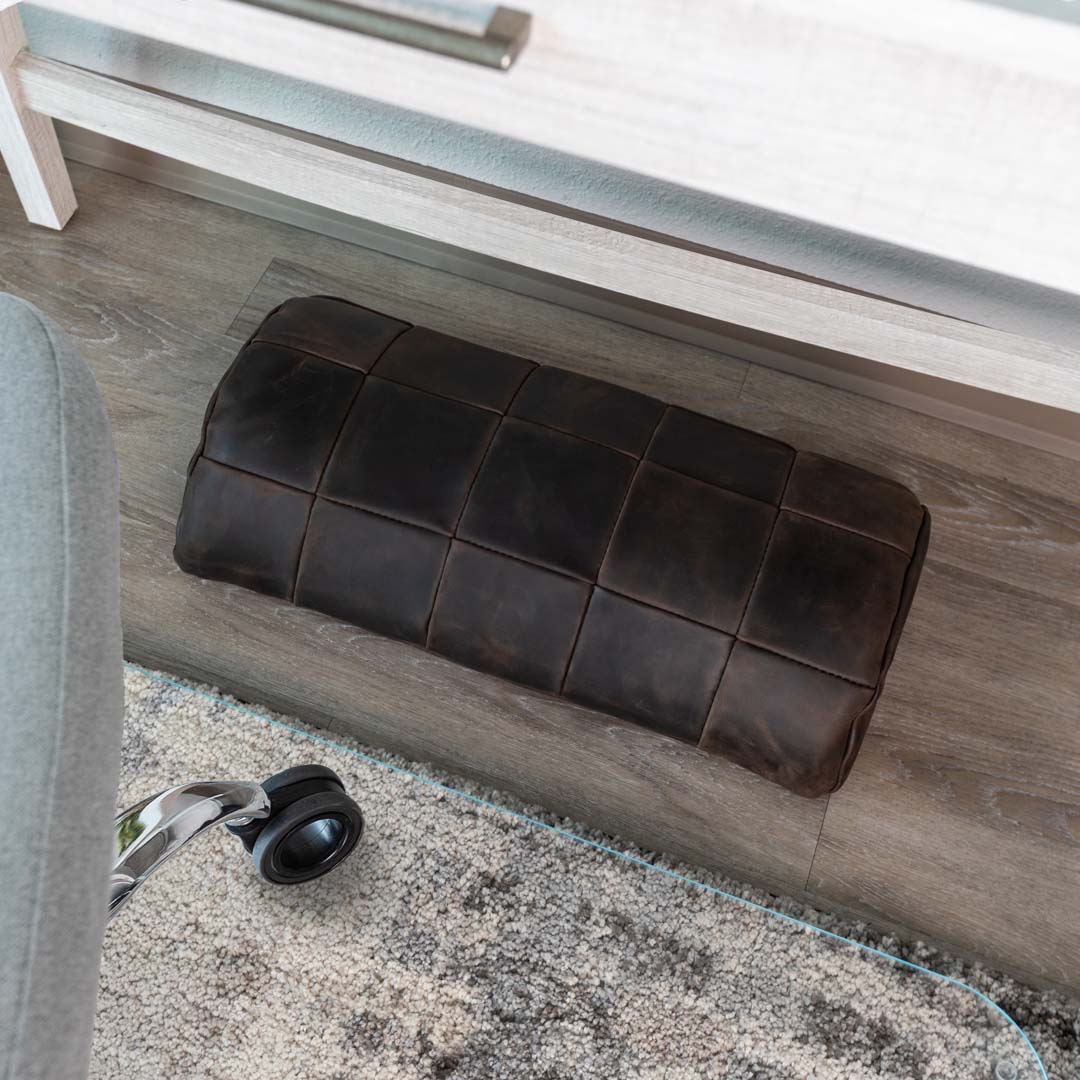 #color_espresso alt="A dark brown, quilted leather foot rest is placed on a light wooden floor, partially resting on a glass chair mat. In the foreground, part of an office chair wheel is visible, and there is a light gray rug with a subtle pattern underneath the chair mat. The background includes a white piece of furniture."
