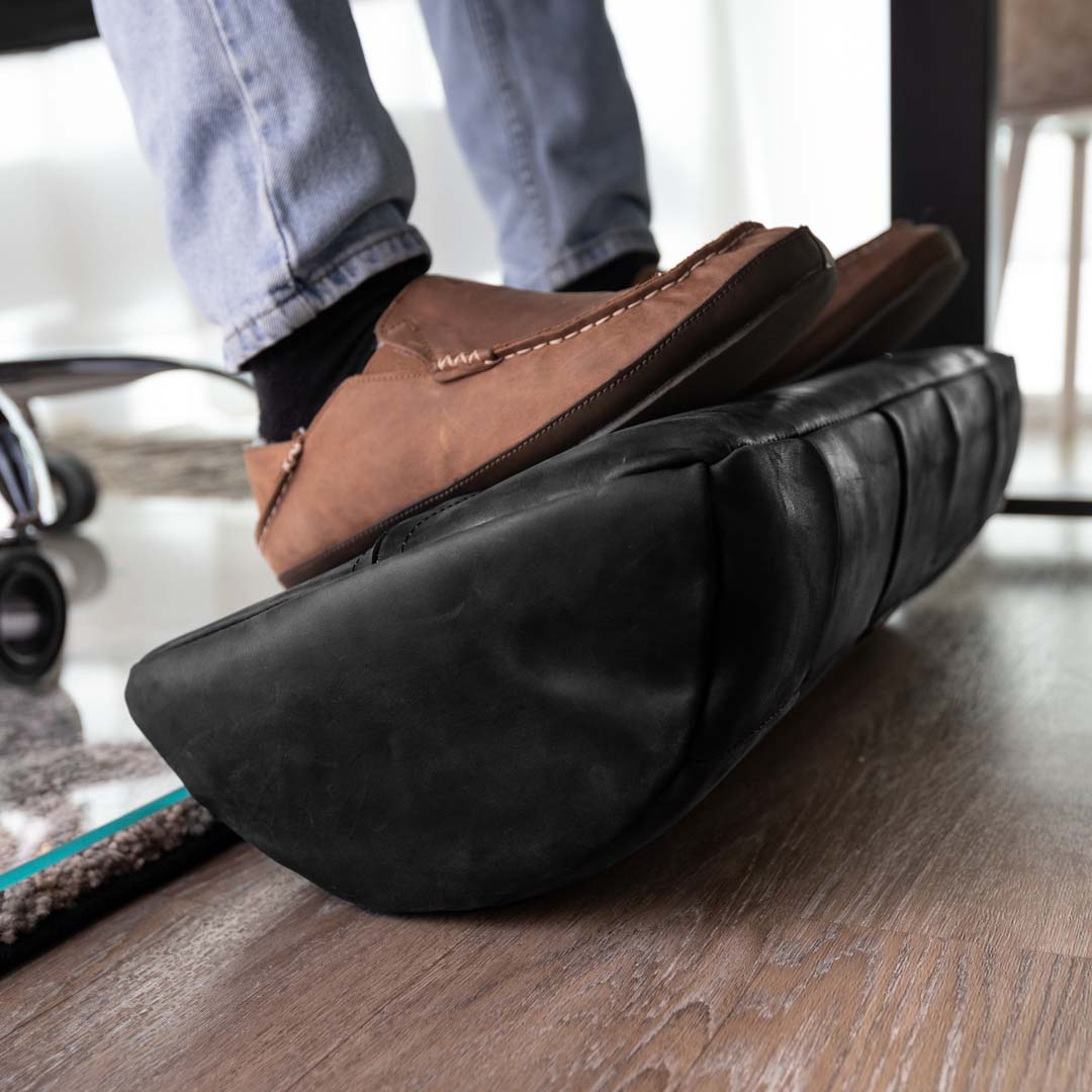 #color_charcoal alt="A person wearing blue jeans and brown leather loafers is resting their feet on a black, quilted leather foot rest. The foot rest is placed on a light wooden floor, and part of a glass chair mat is visible in the background."