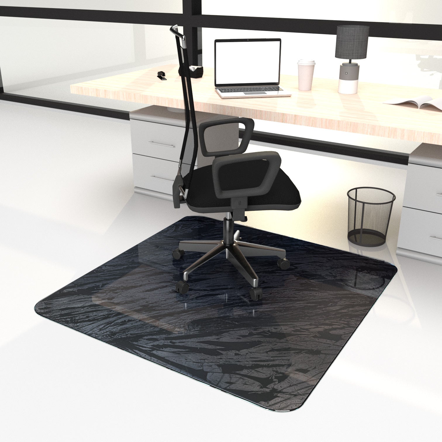 "A modern office setup with a black office chair on a dark, textured chair mat placed on a white floor. The chair is positioned in front of a light wooden desk that has a laptop, a coffee cup, a lamp, and some books on it. The desk also has drawers on either side, and there is a black wire trash bin next to the chair."