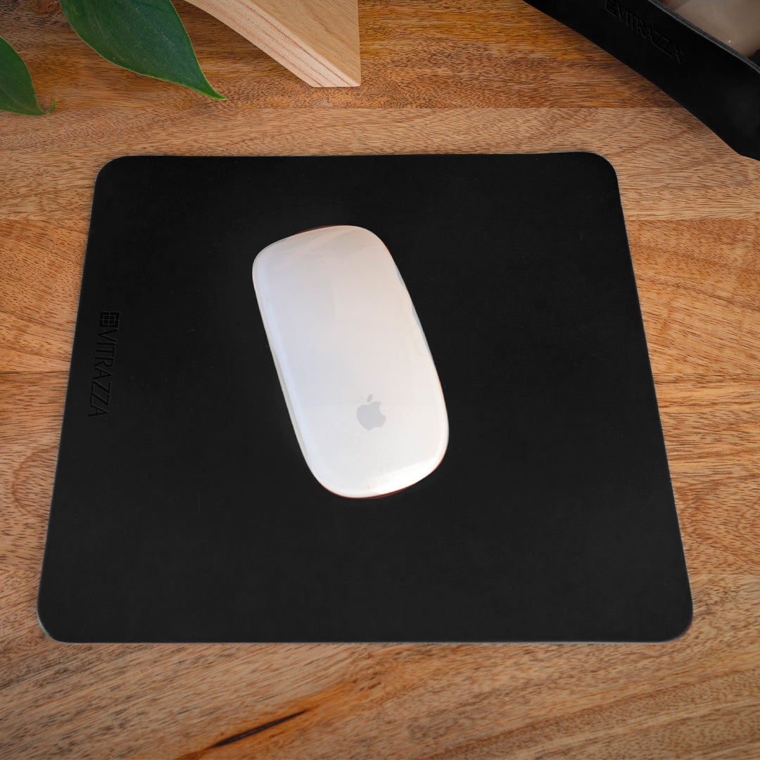 #color_charcoal alt="A white wireless mouse is placed on a black leather mouse pad on a wooden desk. In the background, there is a hint of green foliage and a part of a wooden structure. The mouse pad has the brand name "VITRAZZA" on it.