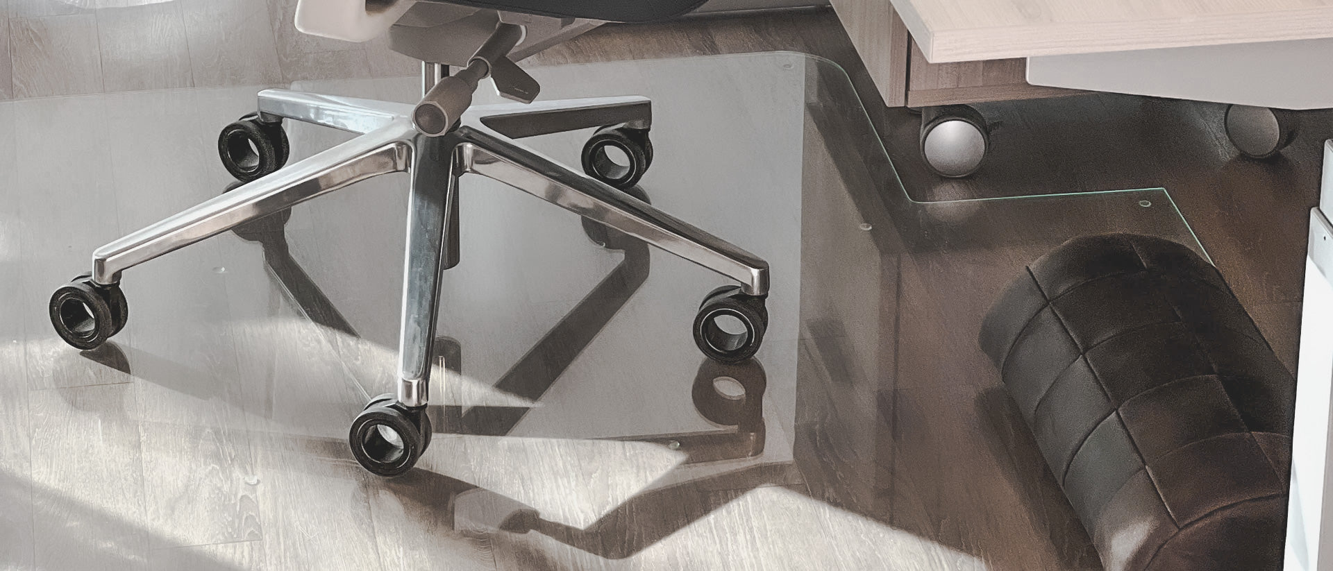 An office chair with a chrome base and black caster wheels is placed on a clear glass chair mat on a wooden floor. To the right, there is a black cushioned footrest and part of a desk with light-colored wood. Shadows of the chair's base are visible on the glass mat.