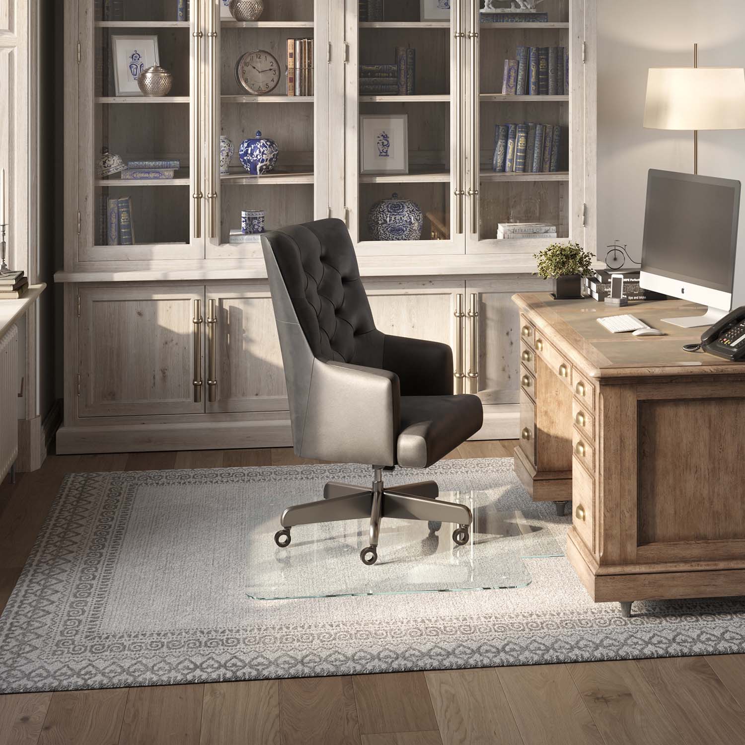 #glass clarity_luminoso (low-iron ultra clear) alt="A home office with a wooden desk, computer, and black leather chair on a 36" x 48" "luminoso" tab glass chair mat over a patterned rug. Behind is a cabinet with glass doors displaying books and decor."
