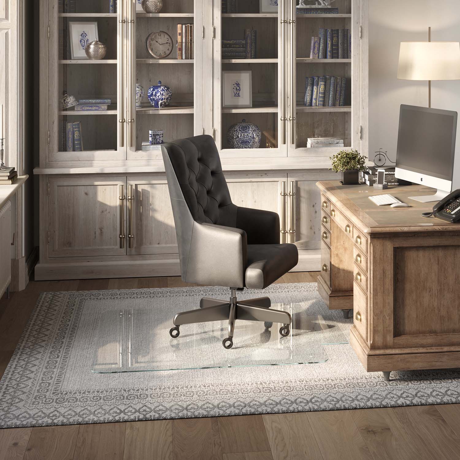 #glass clarity_luminoso (low-iron ultra clear) alt="A home office with a wooden desk, computer, and black leather chair on a 48" x 48" "luminoso" tab glass chair mat over a patterned rug. Behind is a cabinet with glass doors displaying books and decor."
